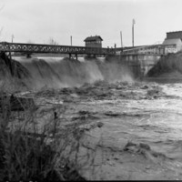 Image: A torrent of flood water pours through a weir along a river. Deep flood waters are visible flowing downstream in the foreground, and a group of men watch the flood from the top of the weir