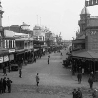 Image: A view from a busy intersection with many people in 1920s attire walking or cycling, and a mix of horse drawn and motor vehicles. The view is centred on a street lined with 2 and 3 storey commercial buildings, most with verandahs and/or balconies. 