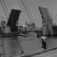 Image: A drawbridge within an active port is open to allow a tug to pass beneath it. A wharf with two mid-twentieth century sailing vessels tied alongside it is visible in the immediate foreground