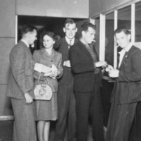 Image: six men and a woman in 1940s era dress stand around a reception desk while a seventh man leans on the desk, writing, behind a glass barrier. In the foreground a second woman in a hat sits looking at the group.