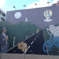 Image: mural of an older man with an ice cream cone watching an abstract figure and a boy both riding bikes on a road in the moonlight. Above, two aliens are on an unidentified flying object