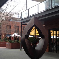 Image: steel quadrangle-shaped sculpture with rounded edges and a geometric hole in the middle in front of a brick courtyard with restaurants' patios