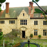 Image: The front of a large, two-storey stone mansion. A garden with benches and statuary is in the middle-foreground
