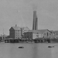 Image: A four-storey stone building is located adjacent to a waterfront wharf. Several ships are visible in the river in the foreground, and two large brick chimneys emerge from behind the stone building