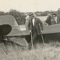 Image: A group of people examine the wreckage of a crashed biplane, including a man using crutches