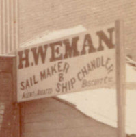 Image: A group of six men of various ages in Victorian attire stand in front of a shop. A painted banner on the top of the shop reads ‘Hy. Weman. Sail Maker & Ship Chandler’