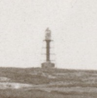 Image: A lighthouse stands at the top of a hill on a desolate island. A low stone building is present next to the lighthouse, and a wooden jetty along the shoreline protrudes into the ocean