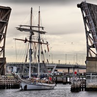 Image: A two-masted sailing vessel passes through a drawbridge on a cloudy morning. Another bridge crossed by modern auto-mobiles is visible in the background