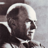 Image: A photographic head-and-shoulders portrait of a balding, middle-aged man in profile. He is wearing a mid-twentieth century suit and reading a document