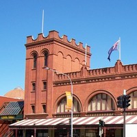 Image: A large brick building with archway windows and a square turret at one end. Cars drive by on a busy street in the foreground