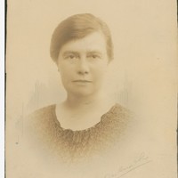 Image: A photographic head-and-shoulders portrait of a woman with ear-length dark hair and wearing 1920s attire