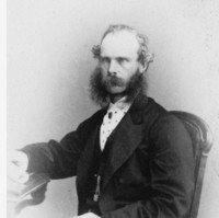 Image: Black and white portrait of a partially balding man with long mutton chops and a moustache sitting in a chair. He is wearing a shirt and suit jacket