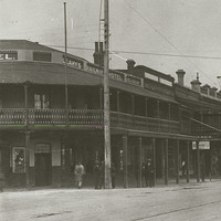 Image: Several Victorian-era buildings line a dirt street criss-crossed by railway tracks. A handful of people and cars of 1920s vintage are visible on the street and adjacent sidewalk