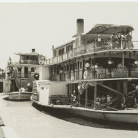 Image: Black and white photograph of two paddlesteamers on a river. Each paddlesteamer has three levels and people are visible standing and sitting on each one. The foremost paddlesteamer is flying the Union Jack flag.