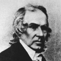 Image: A head-and-shoulders portrait of a middle-aged Caucasian man in mid-nineteenth century clerical attire. He is clean-shaven and has slightly long, shaggy greying hair 