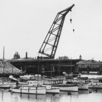 Image: A large derrick crane sits adjacent to one approach to a concrete bridge span. The other half of the bridge, also under construction, is visible in the background