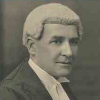 Image: A man in a barrister wig and gown poses for a photograph. He is seated in a chair with his right profile facing the camera