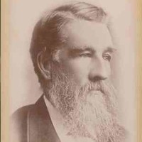 Image: A photographic head-and-shoulders portrait of a middle-aged bearded man in a suit. He has a full head of hair and is looking to the left