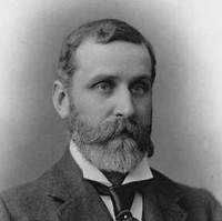 Image: A photographic head-and-shoulders portrait of a bearded, middle-aged man wearing a suit and tie