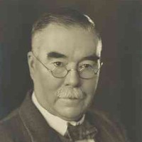 Image: A photographic portrait of a moustachioed middle-aged Caucasian man wearing wire-rimmed spectacles and a mid-20th century suit and tie