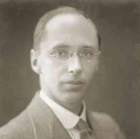 Image: A photographic head-and-shoulders portrait of a young, clean-shaven Caucasian man wearing an early-20th century suit and wire-rimmed spectacles