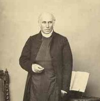 Image: Sepia photograph of bald man standing in front of a chair and table