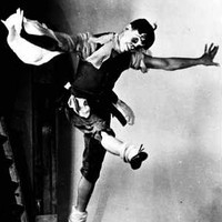 Image: A man in stage make-up and costume dances on a stage with arms outstretched