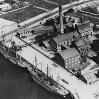 Image: An aerial photograph of a complex of large, multi-storey brick buildings and corrugated metal structures located next to a river. Two sailing ships are moored alongside a wharf associated with the complex