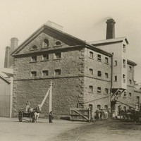 Image: A rectangular five-storey building with peaked roof sits on the edge of a dirt road. People and two horse-drawn carts stand in front of and around the building