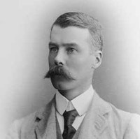 Image: A photographic head-and-shoulders portrait of a young Caucasian man wearing a light-coloured, late-Victorian era suit and sporting a very large handlebar moustache