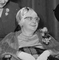 Image: A middle-aged Caucasian woman sits in a wheelchair and is wearing a fur stole and horn-rimmed glasses. Around her are seated three younger Caucasian women in formal dress