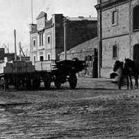 Image: Horse and cart and pedestrians on muddy unpaved roadway with stone and brick buildings either side 