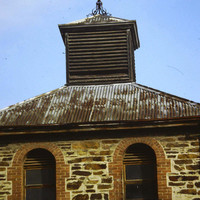 Image: Modern photograph of a bluestone building with arched windows surrounded in red brick and a rusty tin roof, topped with a weather vane.