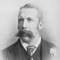 Image: A photographic head-and-shoulders portrait of a young Caucasian man wearing a late-Victorian era suit and sporting a very large handlebar moustache