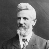 Image: A photographic portrait of a middle-aged Caucasian man with a full beard and head of hair. He is wearing an Edwardian-era suit and tie 