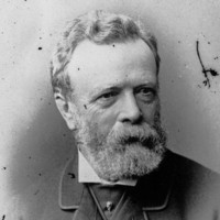Image: A photographic head-and-shoulders portrait of a bearded, middle-aged man wearing a suit with cravat and jewelled cravat pin