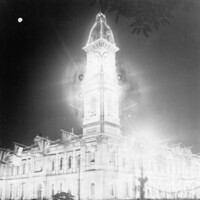 Image: A large, two-storey building with a tall clock tower in one corner is brilliantly lit at night. A small number of people on the street below look at the building