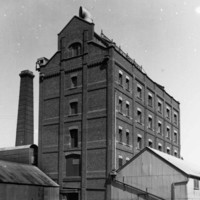 Image: A large, six-storey brick building with several windows on each floor and a large ventilation intake on its roof. Corrugated metal sheds stand in front and to the right of the building