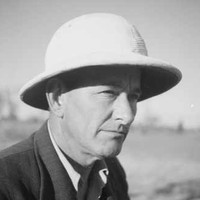 Image: A photographic head-and-shoulders portrait of a middle-aged man wearing a suit-jacket, open-necked shirt and pith helmet
