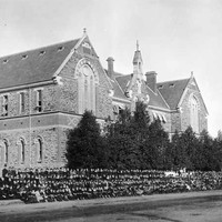Image: Hundreds of schools students in black uniforms pose in lines outside a large symmetrical stone building with decorative brickwork between floors, two huge arched windows on the front gable ends and smaller windows on the sides.
