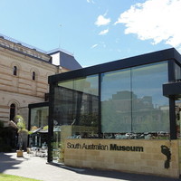 Image: The front of a modern, glass building with a multi-storey historic building in the background
