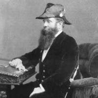 Image: a bearded man in three quarter profile and wearing a hat and sword sits at his writing desk. 