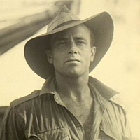 Image: Upper body portrait photograph of a man wearing a shirt with sleeves rolled up, and a large wide brimmed hat