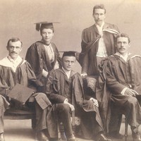 Image: Four men and a woman in university graduation attire sit on chairs and pose for a photograph
