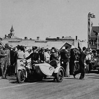 Image: A procession of 1940s-era cars escorted by a policeman on a motorcycle travels through a crowd of people gathered on the approach to a bridge