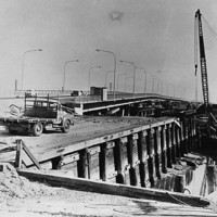 Image: Large cranes dismantle an old bridge span over a narrow river. Cars drive across a new bridge located a short distance and parallel to the old one