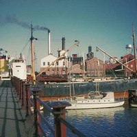 Image: A modern steel-hulled vessel passes through the centre of an historic swing bridge. Another modern boat is moored next to the bridge, and a large complex of historic industrial buildings are visible in the background