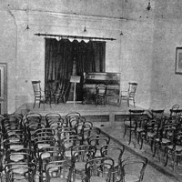 Image: A large, open hall with a stage at one end. Several rows of chairs face the stage, which features additional chairs, a music stand, and a piano