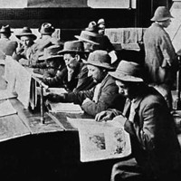 Image: A group of men in hats sit at two long tables and read newspapers