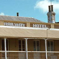 Image: Colour photograph of a two-storey late Victorian-era building on the corner of two paved streets. The building is painted tan with white columns and has the words ‘Railway Hotel, 1849’ painted beneath its roof  